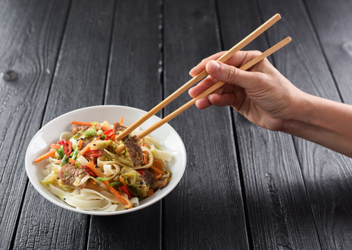 Slender woman hand with chopsticks reaching for Asian udon noodles with meat and vegetables on black background copyspace. Minimalist style