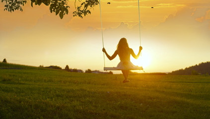 SILHOUETTE: Unrecognizable girl swinging on a tree swing at golden summer sunset - obrazy, fototapety, plakaty