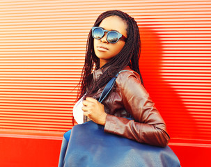 Stylish african woman in sunglasses with bag walking at city over red background