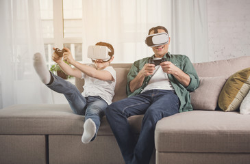 Joyful father and son are playing video game and laughing. They are using virtual reality headsets while sitting on sofa at home - Powered by Adobe