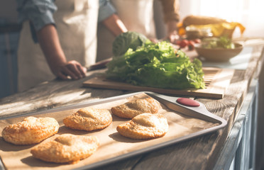 Focus on tasty baked buns on tray. Mature couple is standing at table on background and preparing dinner