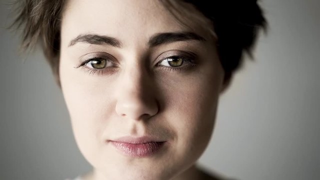 Beautiful young woman with short brown hair wearing a pink t shirt making a kiss motion with her lips and smilng while standing against a gray background. Locked down slow motion close up shot
