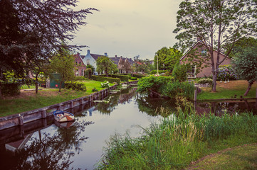 Charming canal next to rustic houses with gardens, trees and boat in cloudy day at Drimmelen. A lovely small hamlet with harbor and elegance streets. Southern Netherlands. Retro vintage filter.