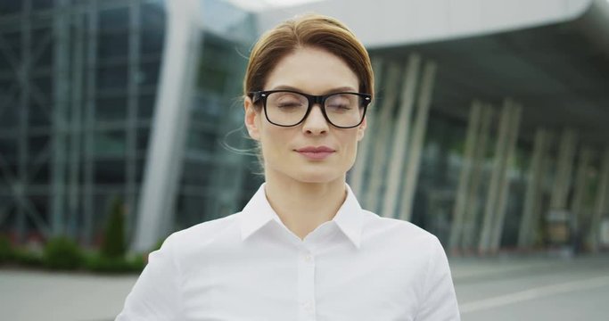Portrait of the pretty Caucasian business lady in glasses looking straight into the camera and smiling on the huge urban glass building background. Close up. Outdoor