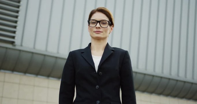 Portrait shot of the Caucasian pretty business lady in the glasses and a black jacket smiling to the camera while standing on the huge glass building background. Outdoor