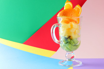 Pieces of tropical fruits in a glass, fruit cocktail. Sliced orange, kiwi cubes, slices of pineapple in a glass bowl. Multi-colored pop art background