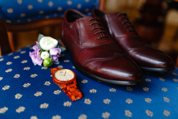 A men's set of accessories on an old wooden chair with a soft seat. The concept of business accessories in clothes. Stylish wristwatch, wooden watch. Men's classic leather shoes, flower boutonniere.