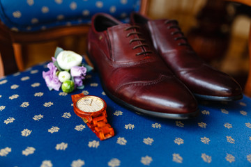 A men's set of accessories on an old wooden chair with a soft seat. The concept of business accessories in clothes. Stylish wristwatch, wooden watch. Men's classic leather shoes, flower boutonniere.
