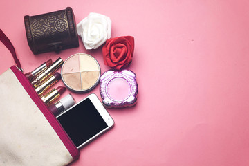 makeup bag contain a differents colorful lipsticks and face powder and phone, jewelry box with red and white flowers  on pink background