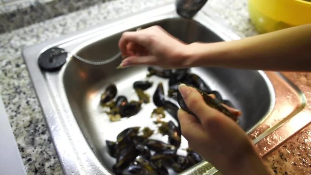 Female hands trying to take off byssus from mussels under the running water in sink. Seafood preparation process.