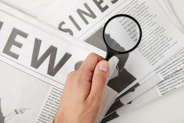 partial top view of person holding magnifying glass above newspapers