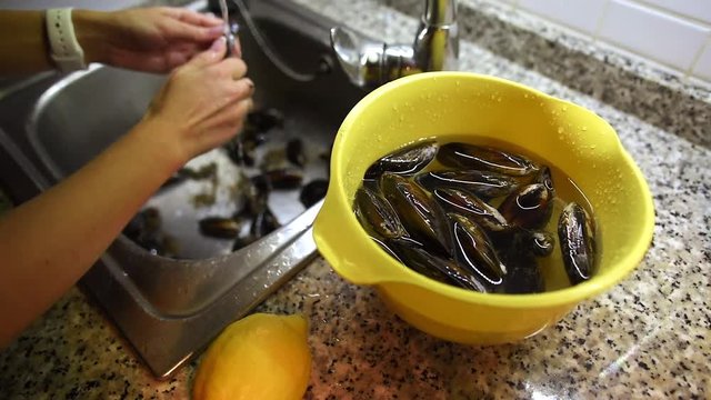 Woman hands taking off byssus from mussels under the running water in sink and then putting them into bowl with water. Seafood preparation process.