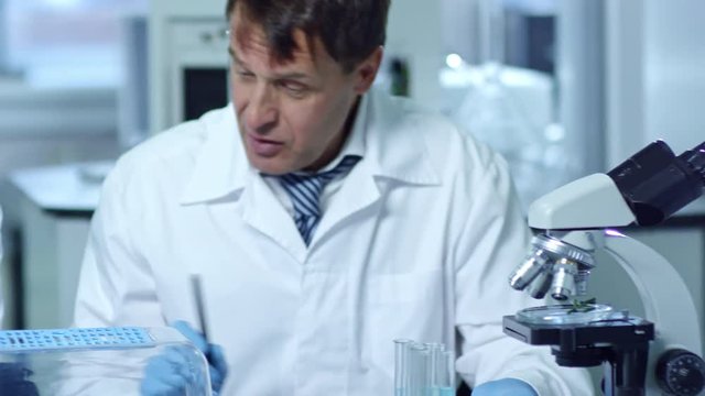 Tracking shot of group of three chemists working in laboratory and studying chemical reactions, one adding drops of liquid to plant specimen and studying it with microscope