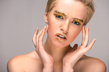 attractive irritated woman with golden glitter on face looking at camera isolated on grey