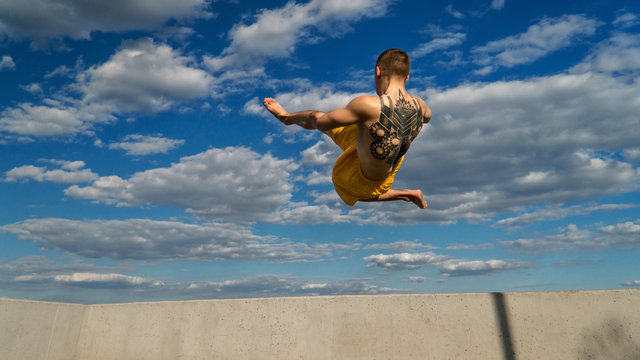 Tricking on street. Martial arts. Man makes high jump barefoot. Shooted from bottom foreshortening against sky.