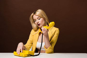pensive blonde girl holding yellow vintage telephone while sitting on brown