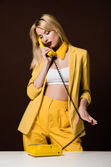 stylish blonde girl in yellow clothes talking by rotary phone on brown