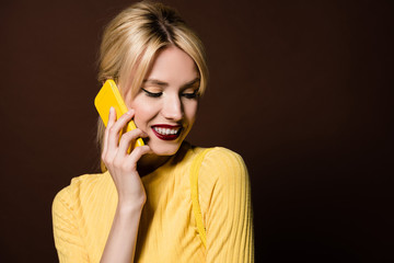 beautiful smiling blonde girl talking by yellow smartphone isolated on brown