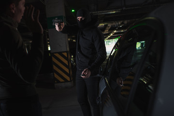 Obraz na płótnie Canvas male mugger in balaclava aiming by gun on man standing near his car