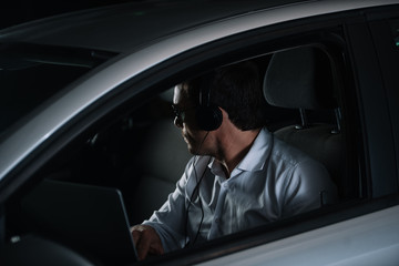 side view of male private detective in headphones doing surveillance with laptop in car