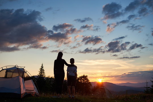 Back view silhouettes of woman hugging child near a camping in the mountains at dawn standing on the grass with wildflowers. The family enjoys the rising sun above the mountains and hills