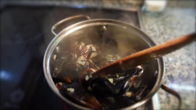 Female hands stirring mussels with wooden spatula in the pan with white sauce of fried onion, garlic and wine. Seafood cooking.