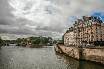 Fototapeta premium Old buildings, wall on the banks of the Seine River and bridges with cloudy sky in Paris. Known as the “City of Light”, is one of the most impressive world’s cultural center. Northern France.