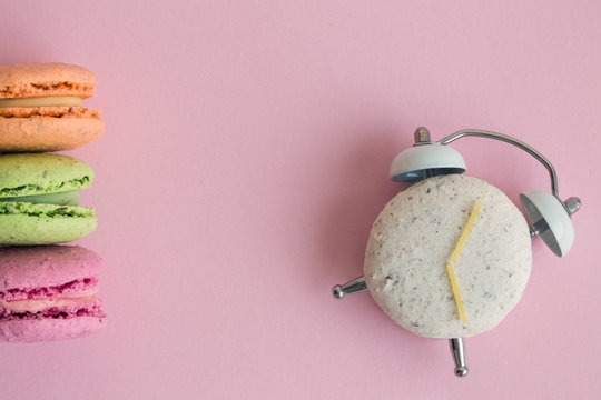 Flat lay of colorful macarons on pastel pink background. One of them in shape of alarm clock with clock hands made of spaghetti.