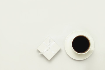 White cup with black coffee, white gift box with white ribbons on a white background. Flat lay, top view
