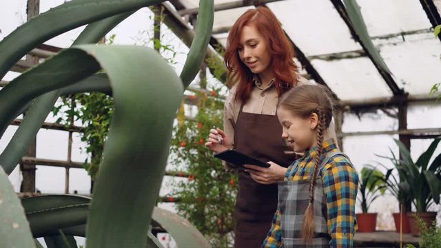 Experienced female gardener and her cute daughter are looking at plants in greenhouse and talking, caring mother is teaching her curious daughter.