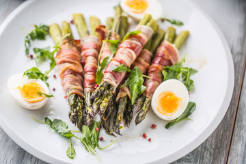 Fresh asparagus wrapped in bacon  on a white plate with arugula tomatoes and eggs
