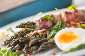Fresh asparagus wrapped in bacon  on a white plate with arugula tomatoes and eggs