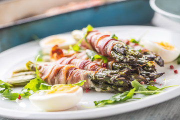 Fresh asparagus wrapped in bacon  on a white plate with arugula tomatoes and eggs