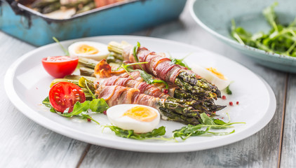 Fresh asparagus wrapped in bacon  on a white plate with arugula tomatoes and eggs