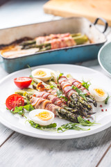 Fresh asparagus wrapped in bacon  on a white plate with arugula tomatoes and eggs