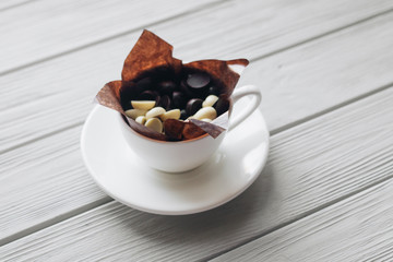 Flat lay of delicious sweet chocolate drops in cup, white wooden background