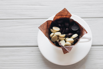 Flat lay of delicious sweet chocolate drops in cup, white wooden background