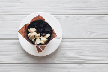 Flat lay of delicious sweet chocolate drops in cup, white wooden background