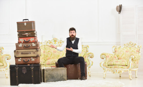 Macho elegant on surprised face sits, confused at end of packing, near pile of vintage suitcases. Luggage and relocation concept. Man with beard and mustache packed luggage, white interior background.
