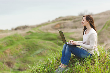 Young smiling successful smart business woman or student in light casual clothes sitting on grass using laptop in field working outdoors on green background. Mobile Office. Lifestyle, leisure concept.