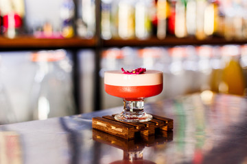 A lifestyle photo of a clover club alcoholic drink in a cocktail glass, served on a small wooden board, standing on a steel barstand. Selective focus, grain effect, bokeh background.