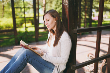 Portrait of beautiful calm peaceful young woman wearing light casual clothes, jeans, relaxing, reading book. Smiling female resting in city park in street outdoors on spring nature. Lifestyle concept.