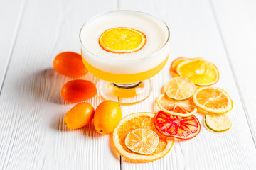 A photo of a bright orange alcoholic drink in a cocktail glass, garnished with dried orange, lime and grapefruit and fresh cumquat. White wooden table.