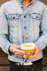 Man in a blue jean jacket holding a bright orange alcoholic drink in a cocktail glass, garnished with dried orange.