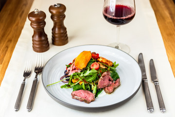 A warm salad with roastbeef, arugula, cherry tomatoes, dried carrot and grilled cheese. Served with a glass of red wine, salt and pepper. Forks and knifes.