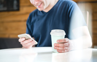 Fototapeta na wymiar Happy man using smartphone in cafe or home. Smiling guy holding mobile phone and paper coffee cup. Texting, shopping online or browsing internet in coffee shop.