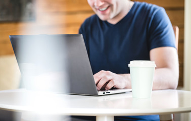 Happy man using laptop with take away coffee cup on table. Smiling person with computer home or in cafe.