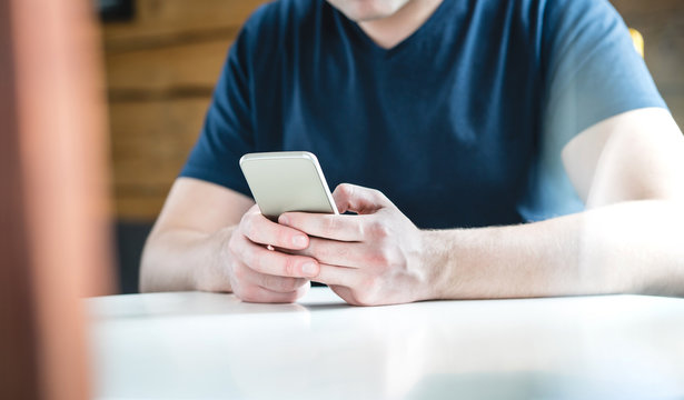 Young man texting with smartphone. Guy using mobile phone against wooden wall and background. Cropped close up shot of hands holding cellphone on table.