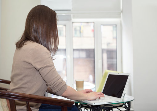 Brunette Woman Working With A Laptop Near A Window. Side View, No Face Visible.