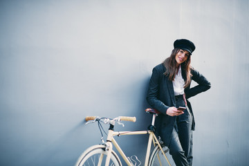 Woman standing against the wall and using smart phone for texting. Next to her yellow bicycle.